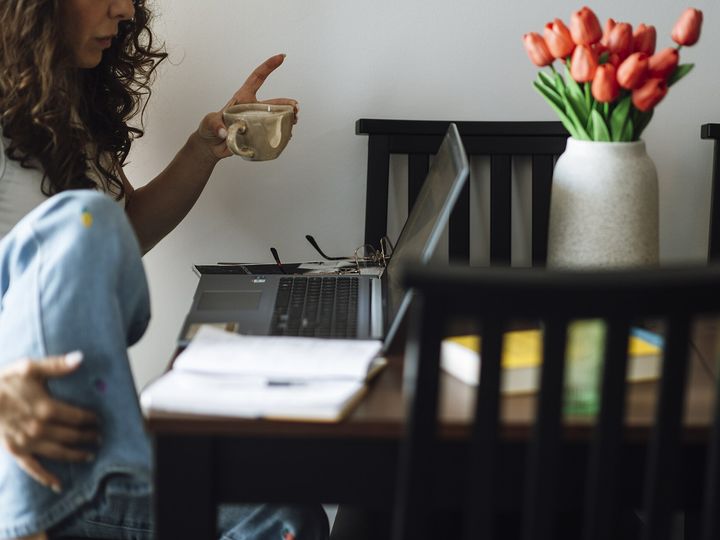 A person standing at a desk with an open laptop, notebooks and a vase of tulips.