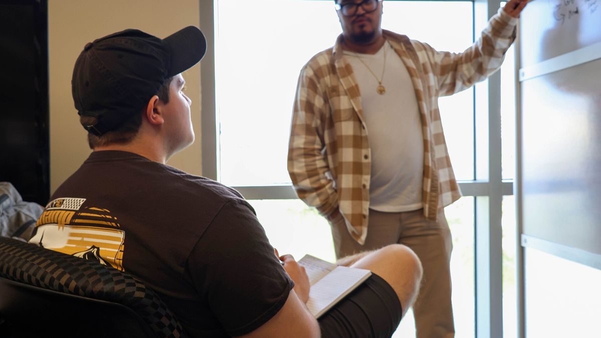 Two people talking across a room, one seated with a clipboard on his knee.